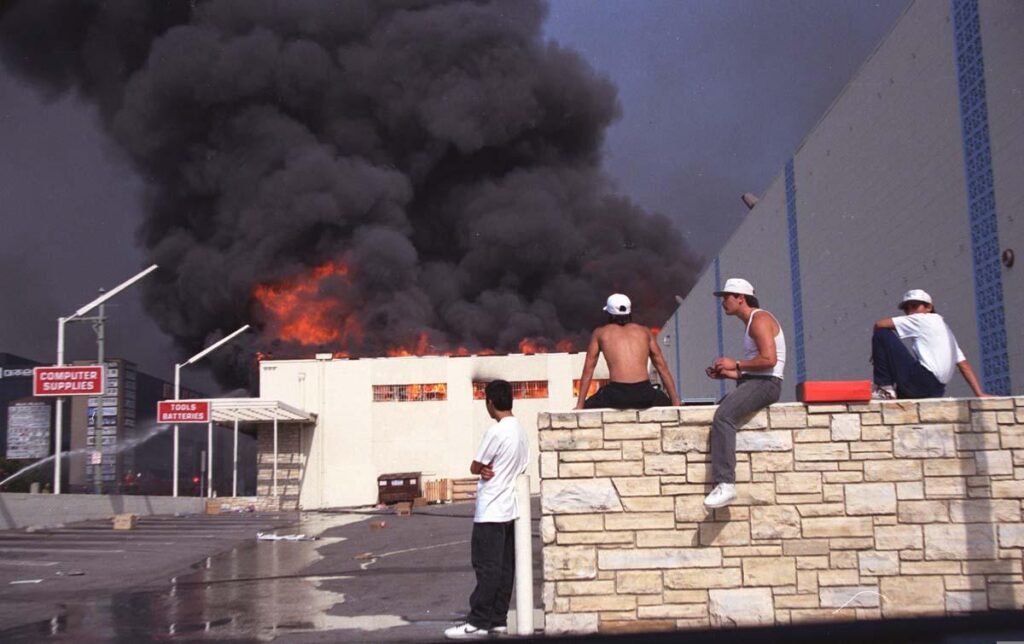 A color photograph of a burning white computer supplies and tools shop in the background. In the foreground a tall stone-lab adorned wall with various men in leaning against or sitting on the wall either looking at hte building or looking at the camera person. 